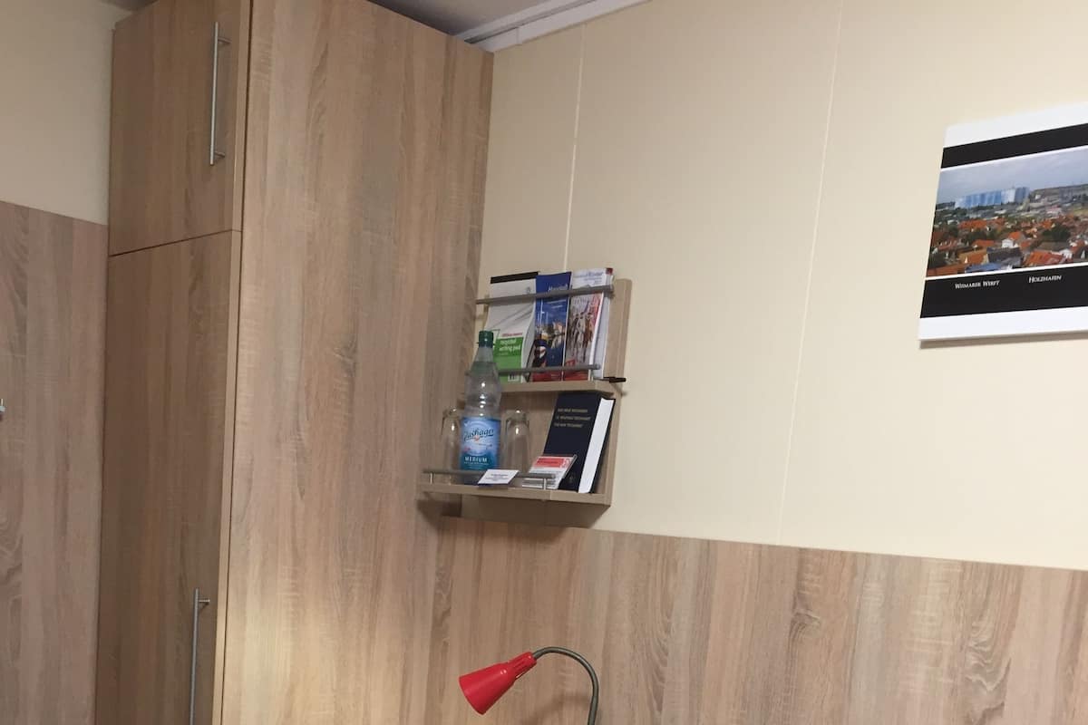 Een kamer met een houten bureau, een rode bureaulamp, een houten stoel, een bankje en een plank met boeken en een waterfles.