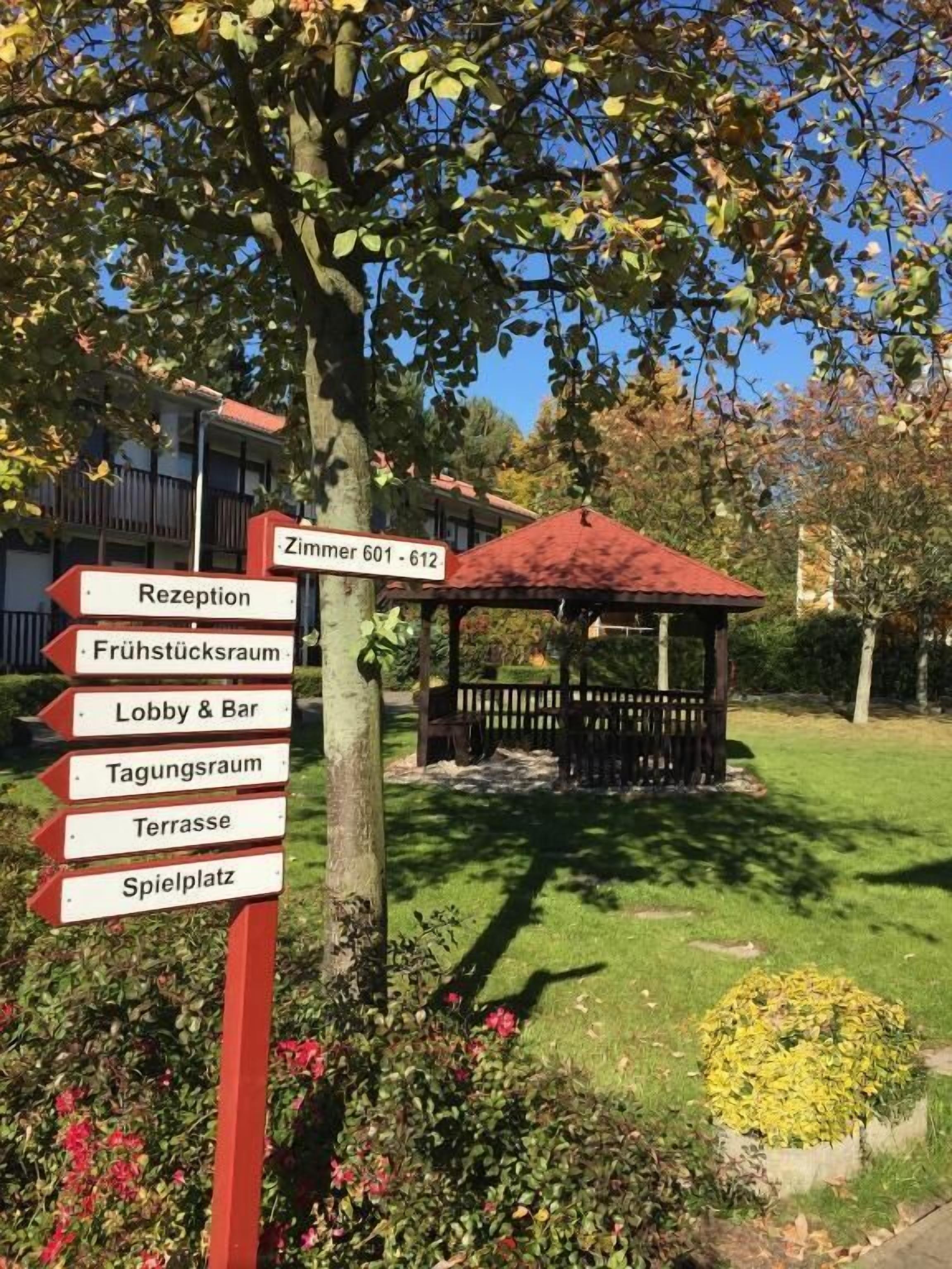 Ein Wegweiser mit Hinweisen zu verschiedenen Bereichen des Hotels, darunter die Rezeption, die Lobby, die Bar, der Speisesaal, die Terrasse und der Spielplatz.