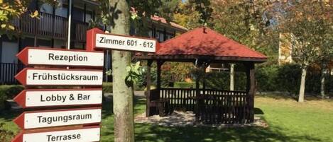 Ein Wegweiser mit Hinweisen zu verschiedenen Bereichen des Hotels, darunter die Rezeption, die Lobby, die Bar, der Speisesaal, die Terrasse und der Spielplatz.