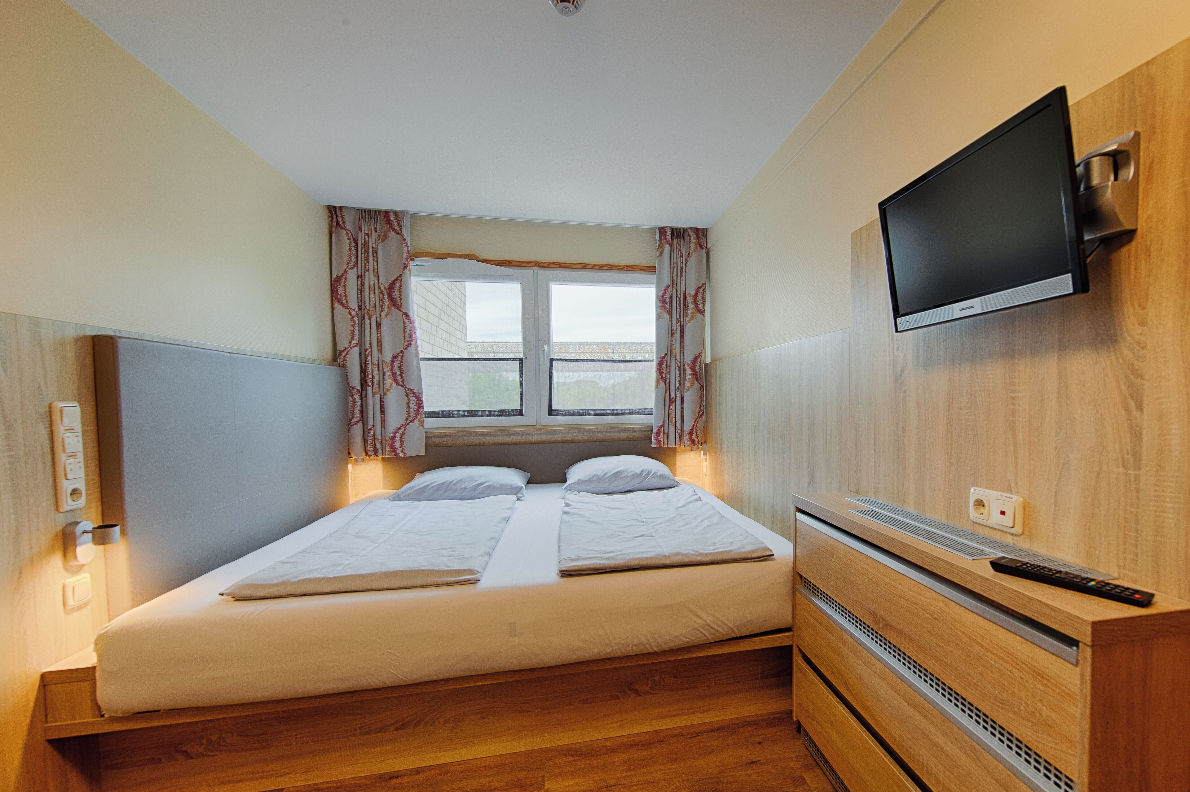 Una habitación de hotel con una cama, un televisor montado en la pared y una ventana con cortinas.
