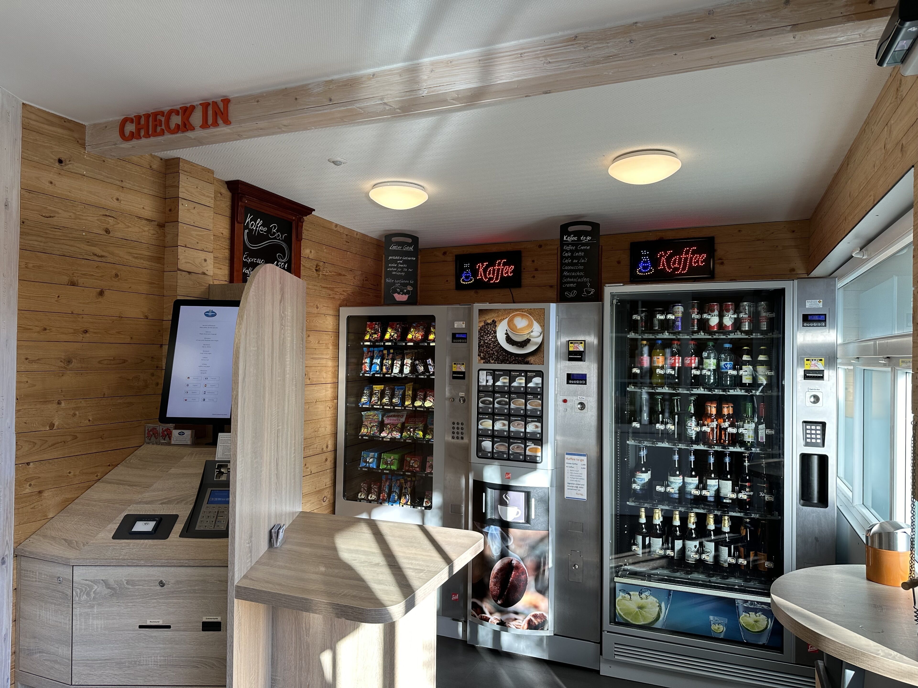 A coffee shop interior with wooden walls, a 'Check In' sign, a digital menu board, and coffee vending machines.