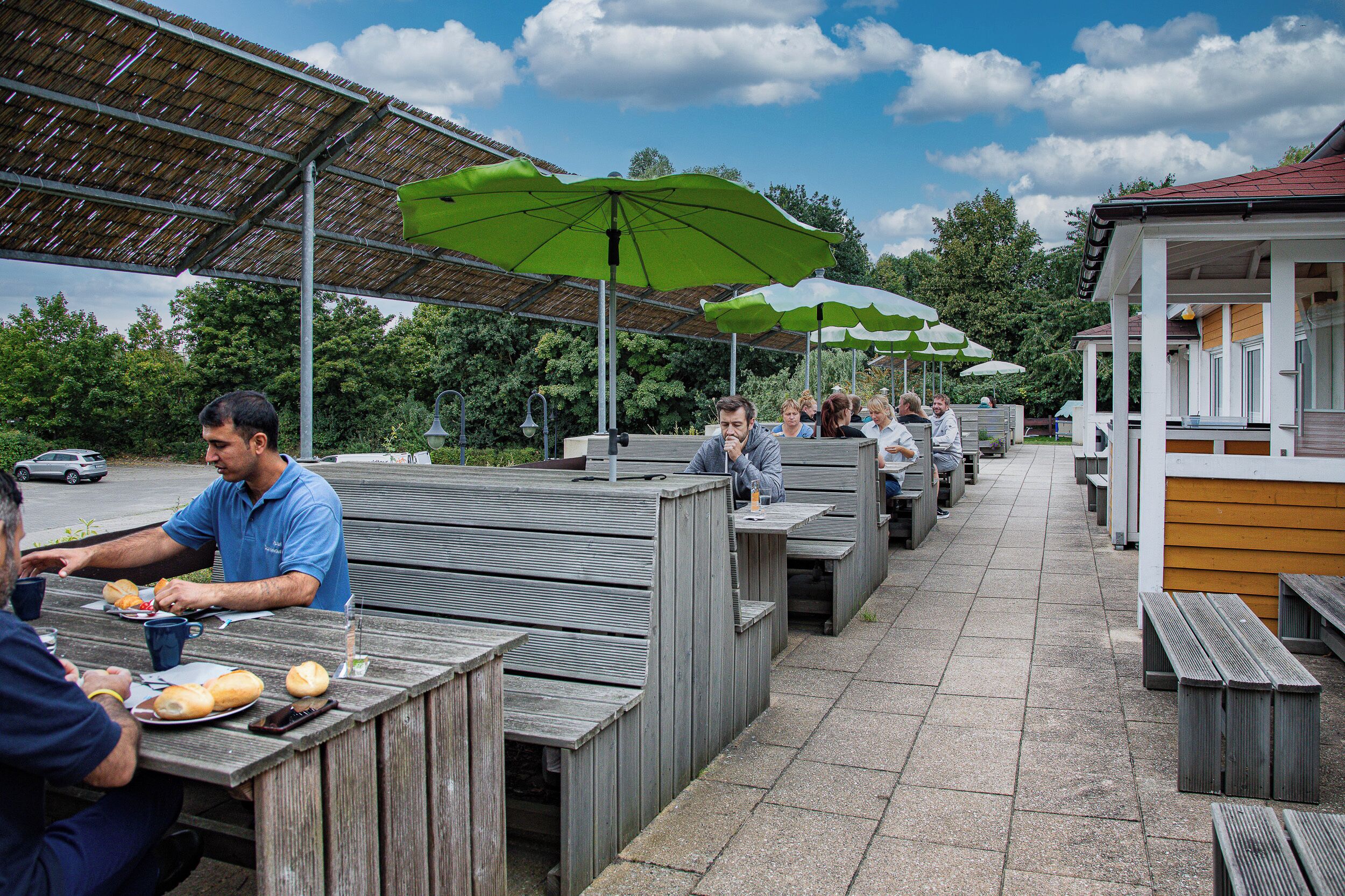 An outdoor dining area with wooden benches and tables, covered by a roof with green umbrellas.