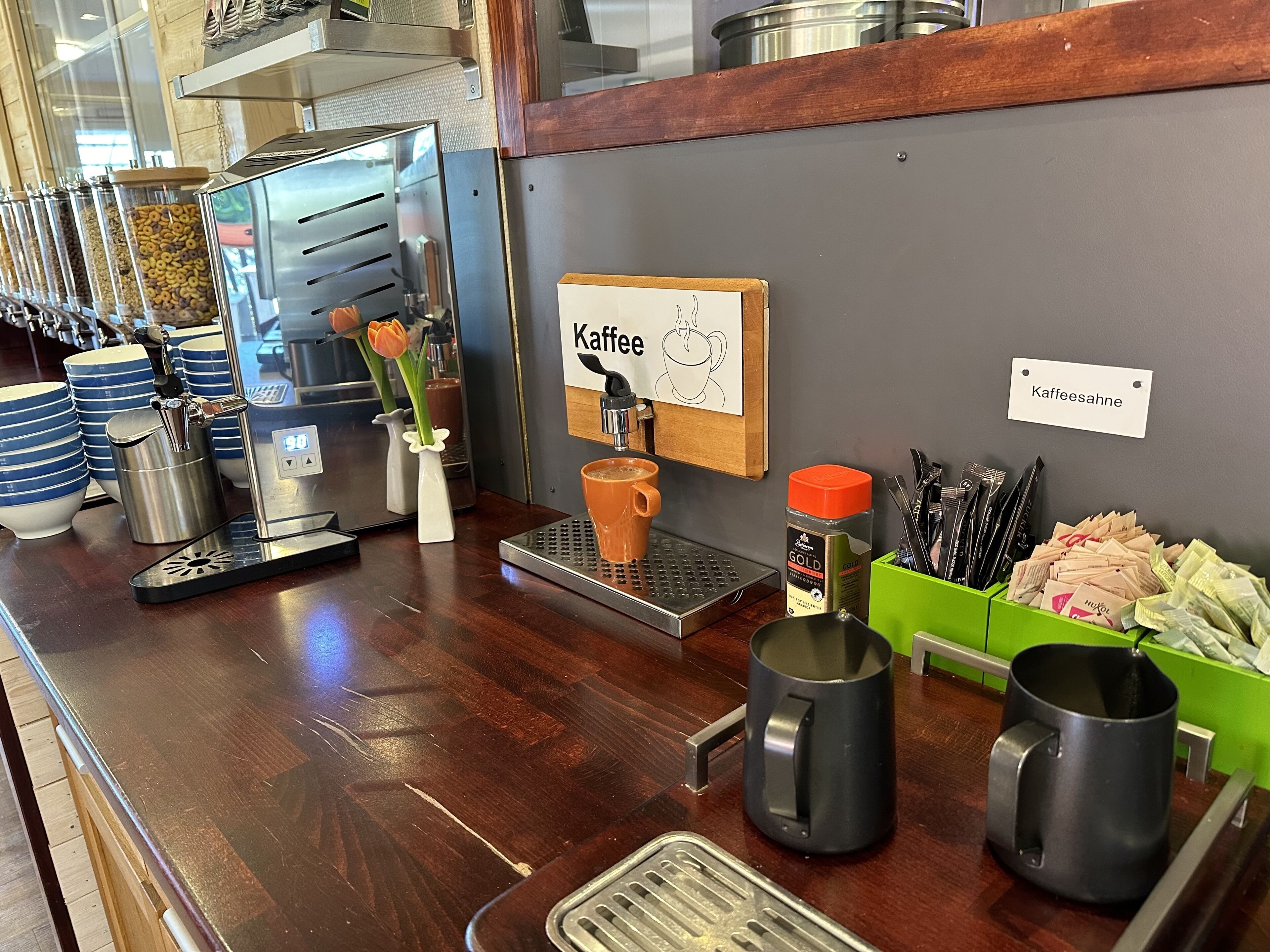Un bar de café con una máquina de café, un cartel que dice "Kaffee" y diversos objetos relacionados con el café.