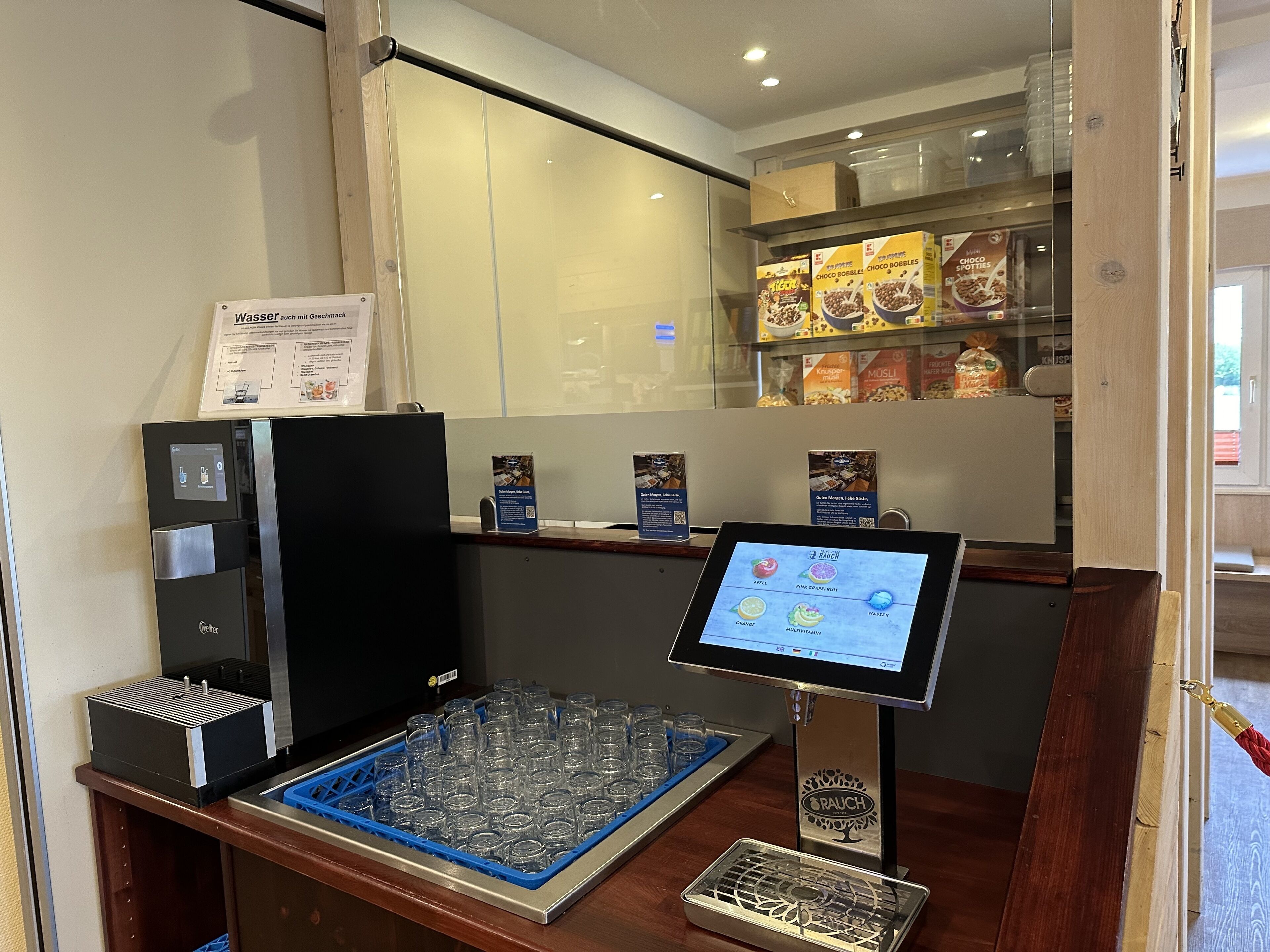 Una estación de bebidas de autoservicio con menú digital, vitrina refrigerada y un porta vasos.