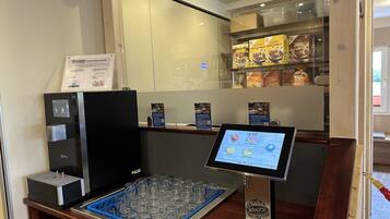 Una estación de bebidas de autoservicio con menú digital, vitrina refrigerada y un porta vasos.