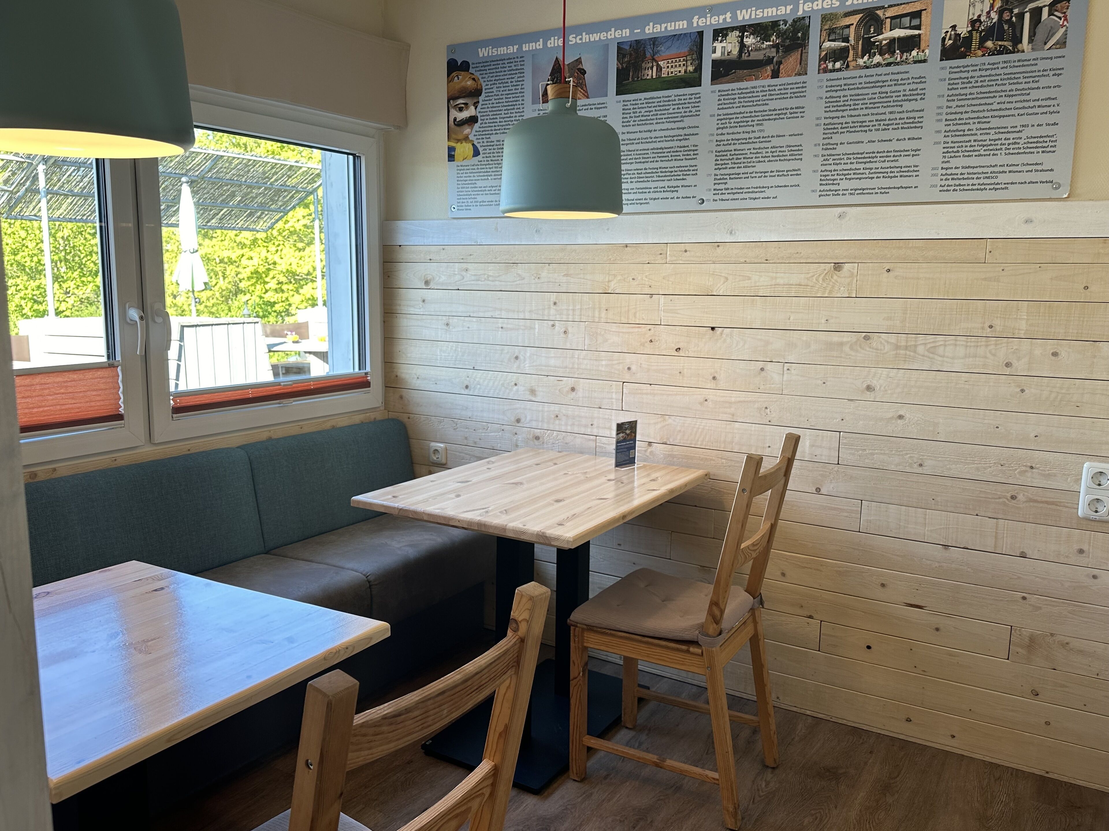 Acogedor interior de cafetería con una mesa de madera y bancos, un ventanal con vistas y un tablero de menú colgado en la pared.