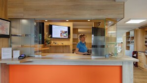 Een receptiebalie met een digitaal scherm waarop hotelinformatie staat, een betaalautomaat en een aanrecht met een houten afwerking.