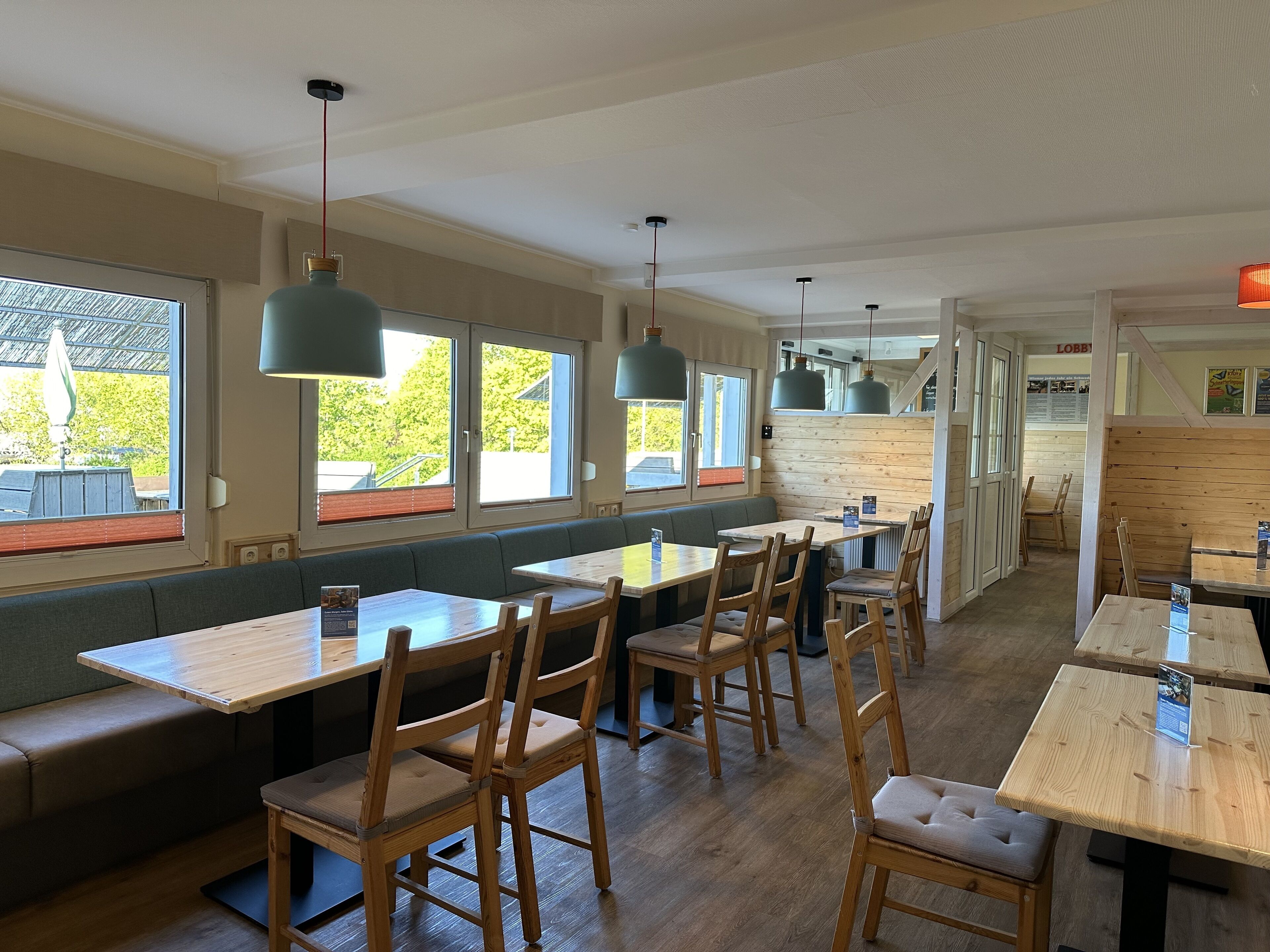 Un restaurante con mesas y bancos de madera, ventanas amplias y lámparas colgantes.