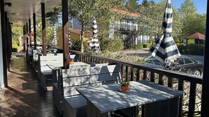 Een terras buiten met houten tafels en gestreepte parasols, met uitzicht op een woonwijk met huizen en een auto.
