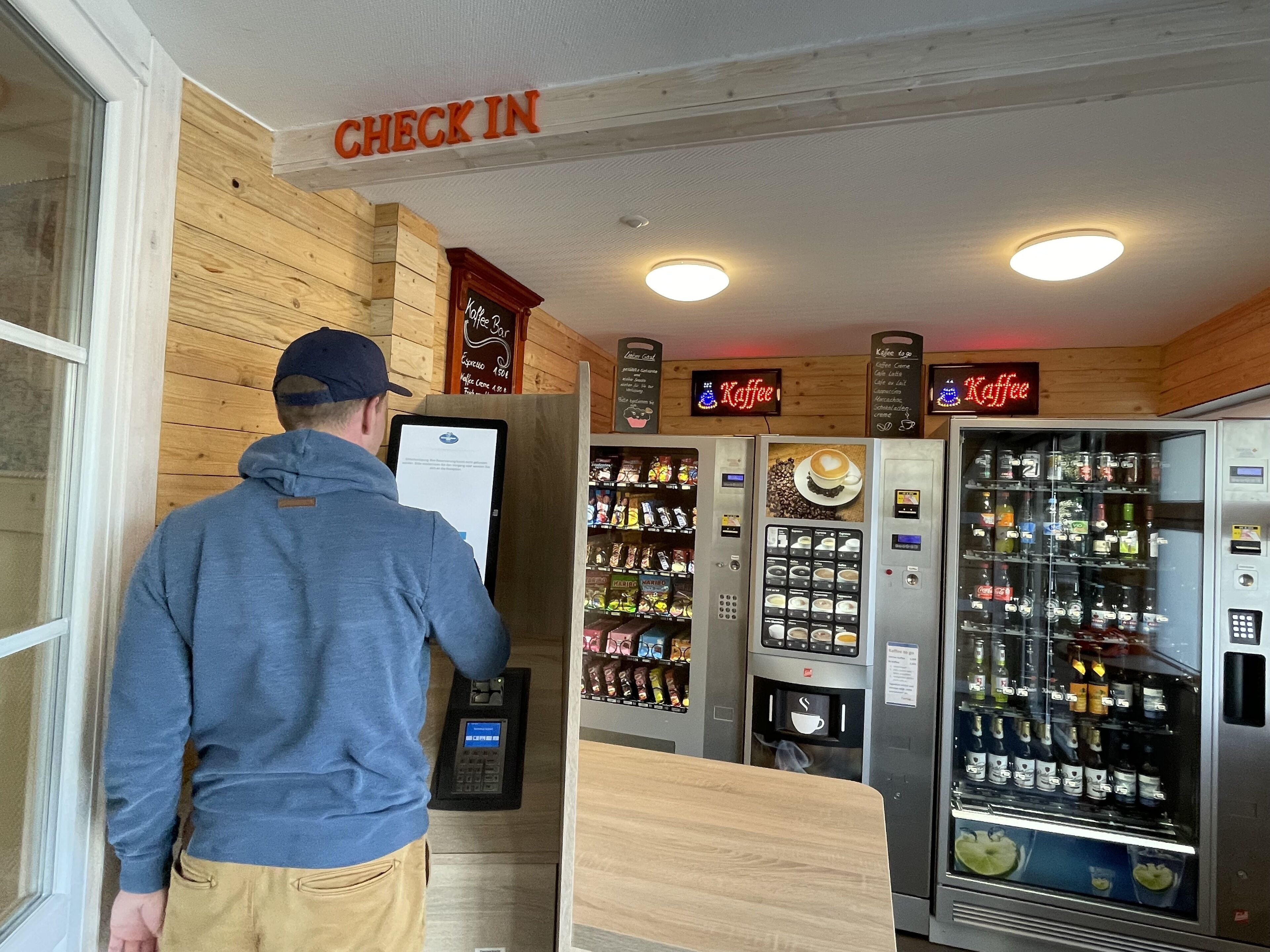 A man at a coffee shop counter with a digital menu and a 'Check In' sign above.