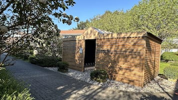 A small wooden structure with a slatted roof, surrounded by a gravel path and greenery.