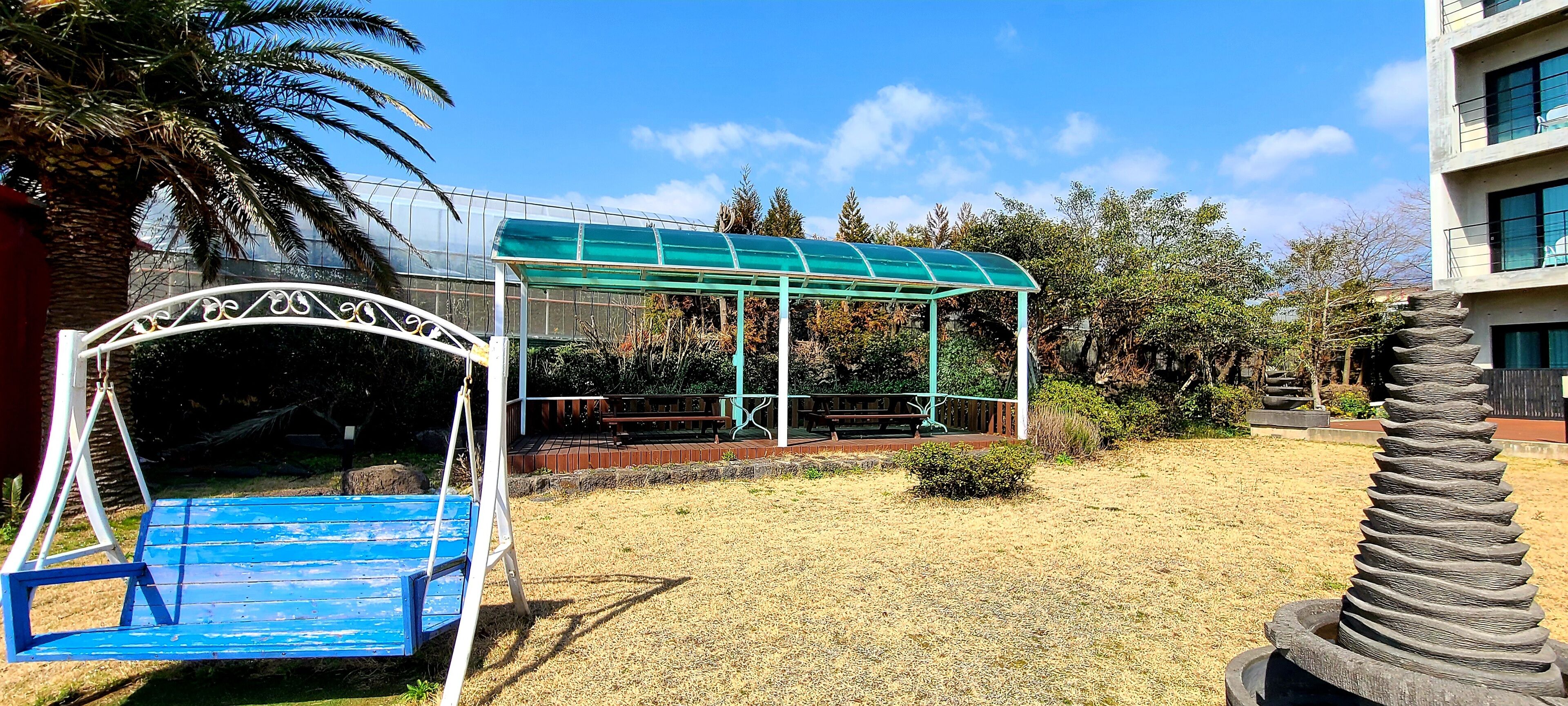 Un espace extérieur aménagé avec des sièges, protégé par un toit transparent, et entouré d’herbe sèche et de palmiers.