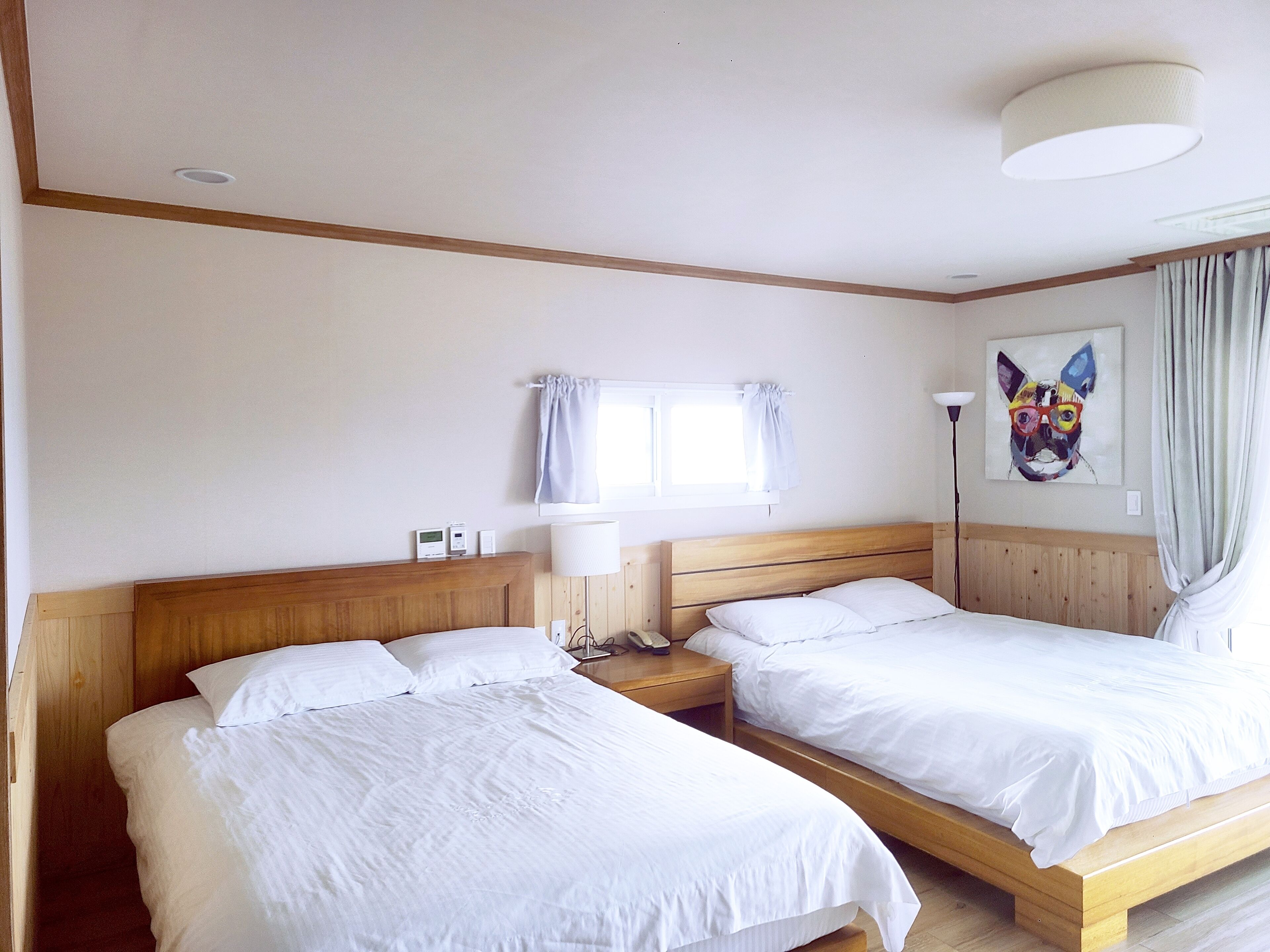 Camera d'albergo con due letti singoli, una testiera in legno, un comodino con lampada, una finestra con tende e un quadro appeso al muro.
