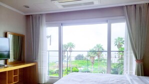 A hotel room with a large bed, a TV on a wooden stand, and a balcony with a view of greenery and buildings.