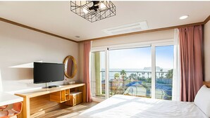 A hotel room with a large bed, a TV, a desk, and a balcony with a view of the sea.
