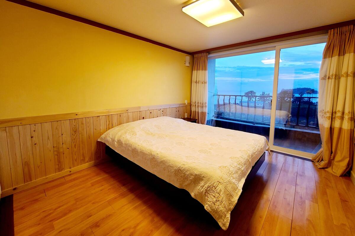 A room with a bed, wooden floor, a window with curtains, and a view of the outdoors.