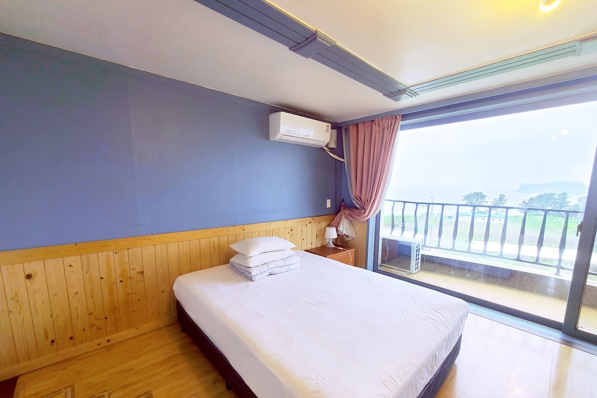 A hotel room with a wooden bed, a nightstand, an air conditioning unit, and a view of the outdoors.