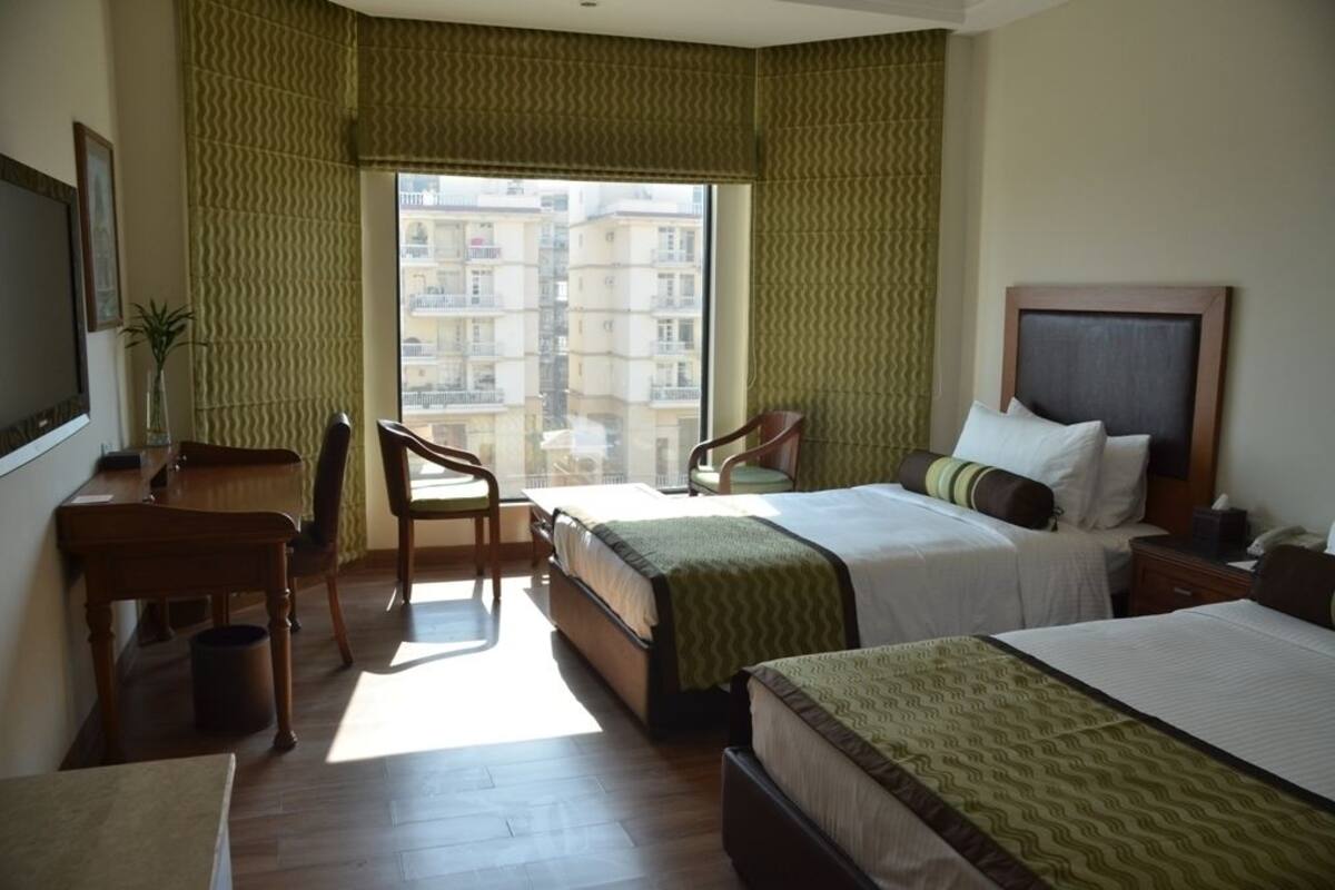 Une chambre d’hôtel avec deux lits, un bureau, une chaise et une grande fenêtre donnant sur des immeubles.
