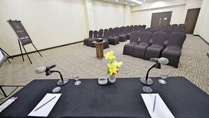 Una sala de conferencias con filas de sillas, una mesa con micrófonos y un caballete.