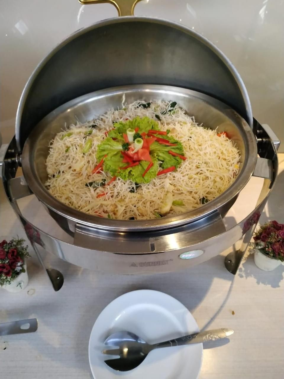 A stainless steel pot with a lid, containing a dish with noodles and vegetables, placed on a table with a spoon and a small white dish nearby.