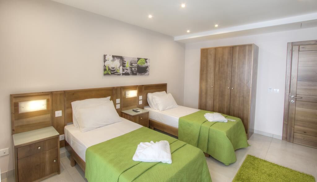 A hotel room with two single beds, wooden headboards, a green rug, a wooden wardrobe, and a painting on the wall.