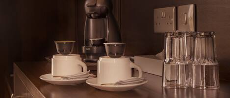 A coffee station with a coffee maker, two cups, two saucers, and three glasses on a wooden counter.