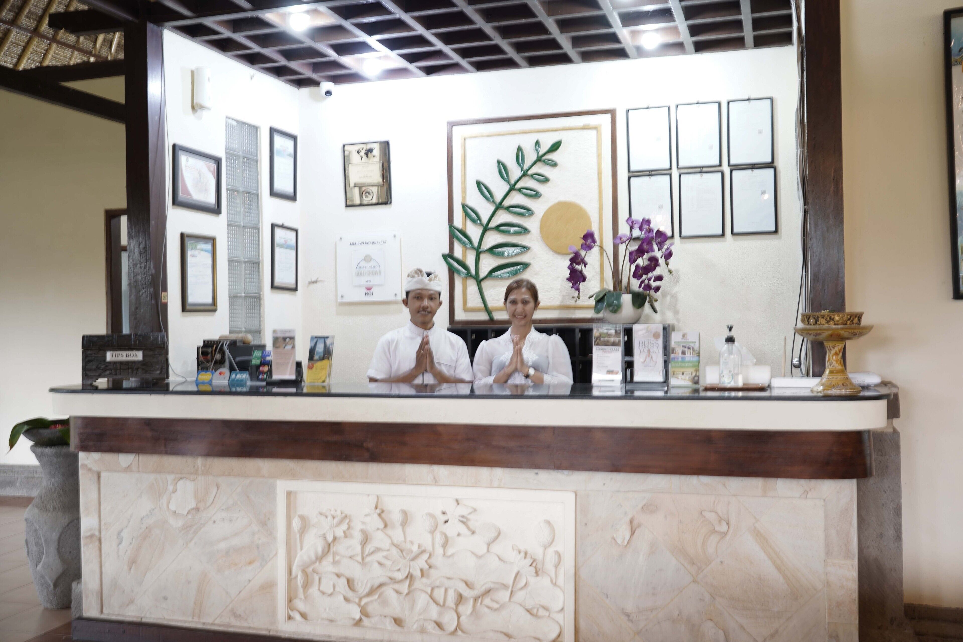 Un mostrador de recepción con certificados enmarcados, una obra de arte de hojas y un jarrón con flores moradas.