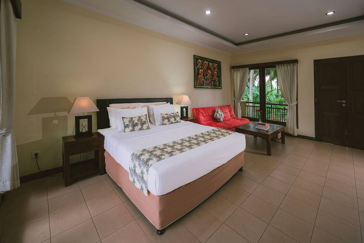 A hotel room with a large bed, two bedside tables with lamps, a red sofa, a small table with books, and a window with curtains.