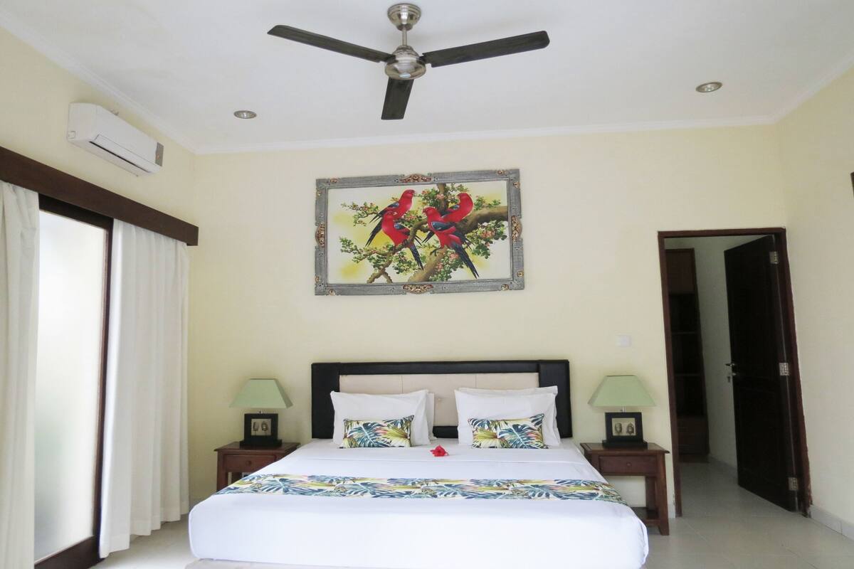 A hotel room with a bed, a ceiling fan, an air conditioning unit, a framed picture of birds, two bedside lamps, and a door leading to another room.