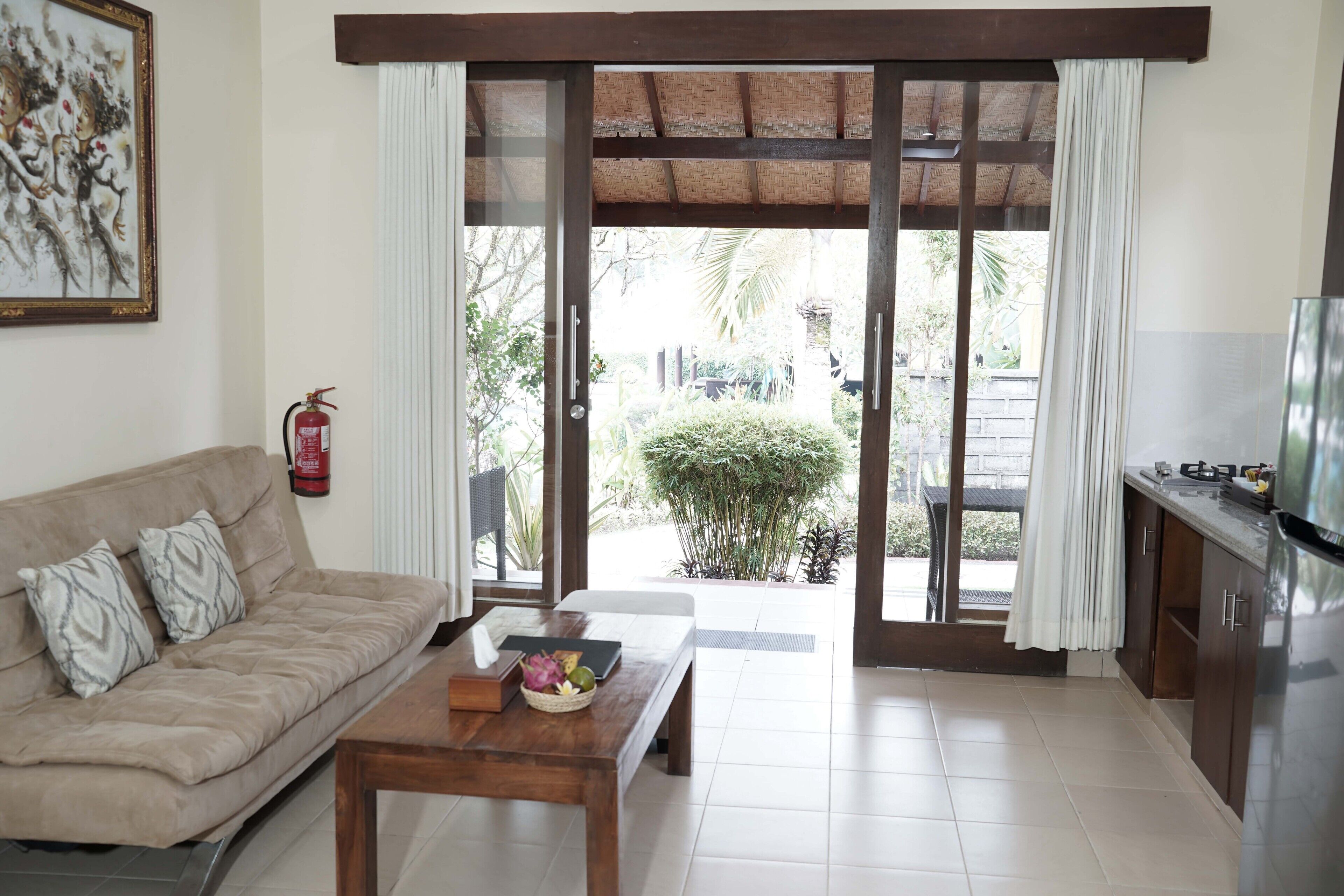 A living room with a sofa, a wooden coffee table, a television, and a sliding glass door leading to a balcony with a view of greenery.