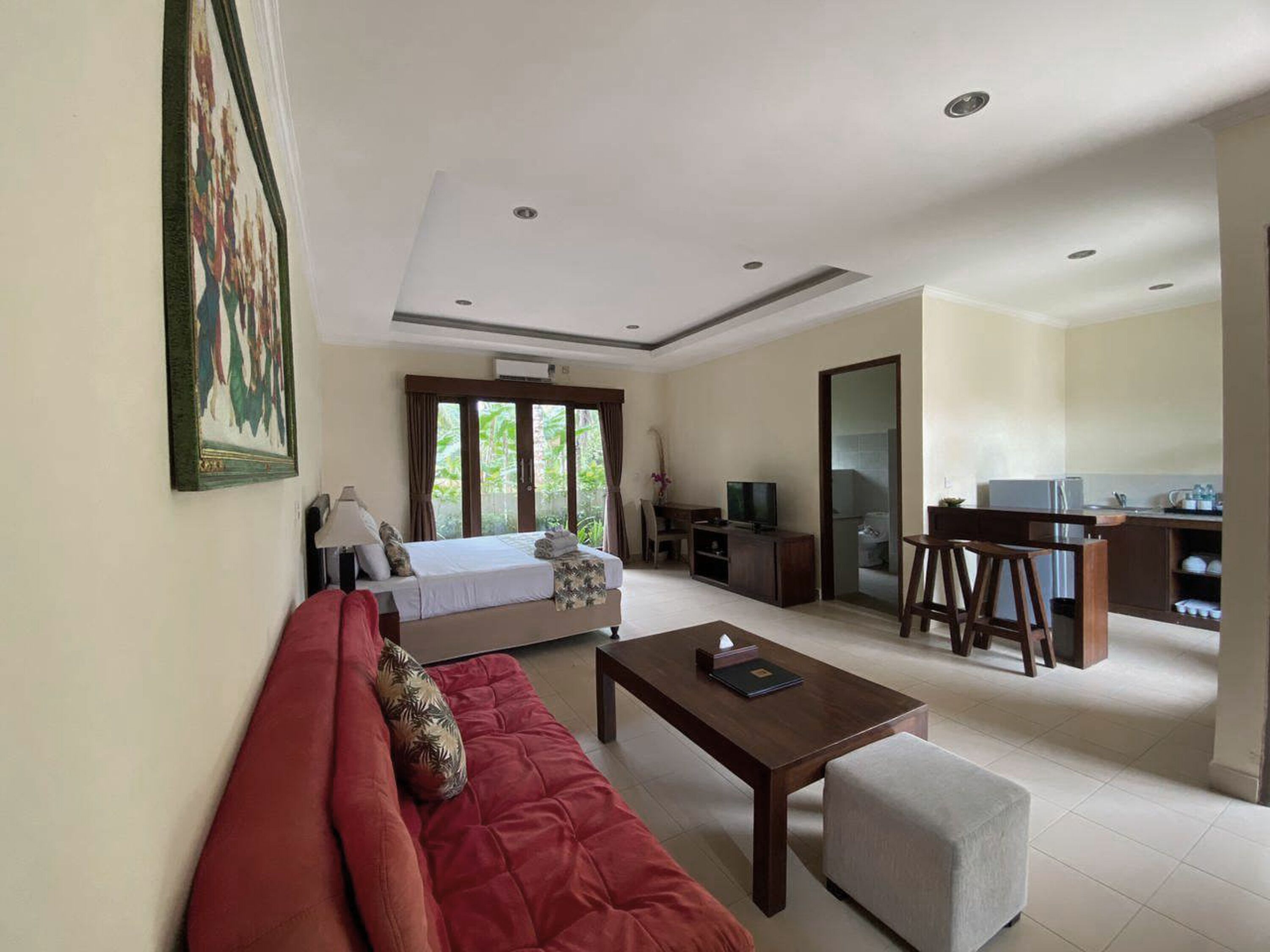 A modern hotel room with a red sofa, a small table, a TV, and a kitchenette in the background.