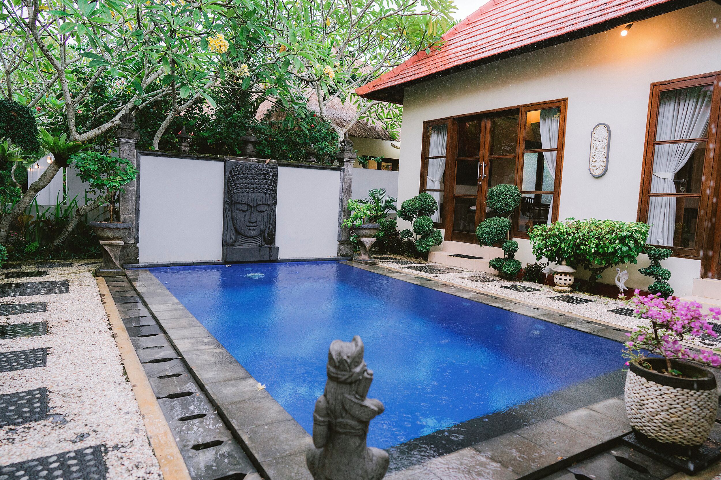A swimming pool with a statue in the foreground, surrounded by potted plants and a house with a red roof.
