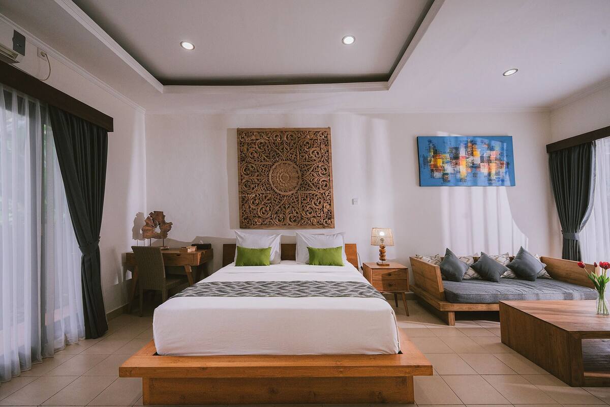 A modern hotel room with a large bed, a sofa, a desk, and a wall art piece.