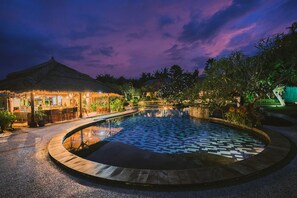 Une piscine circulaire au fond décoré de motifs, entourée d’une zone pavée abritant une structure à toit de chaume et des plantes en pot.