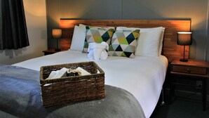 Una cama bien hecha con cabecera, dos mesitas de noche con lámparas, una cesta de mimbre sobre la cama y una manta gris.