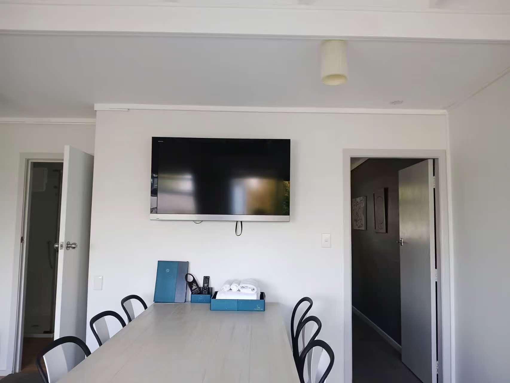 A dining area with a long table, chairs, a flat-screen TV mounted on the wall, and a door leading to another room.