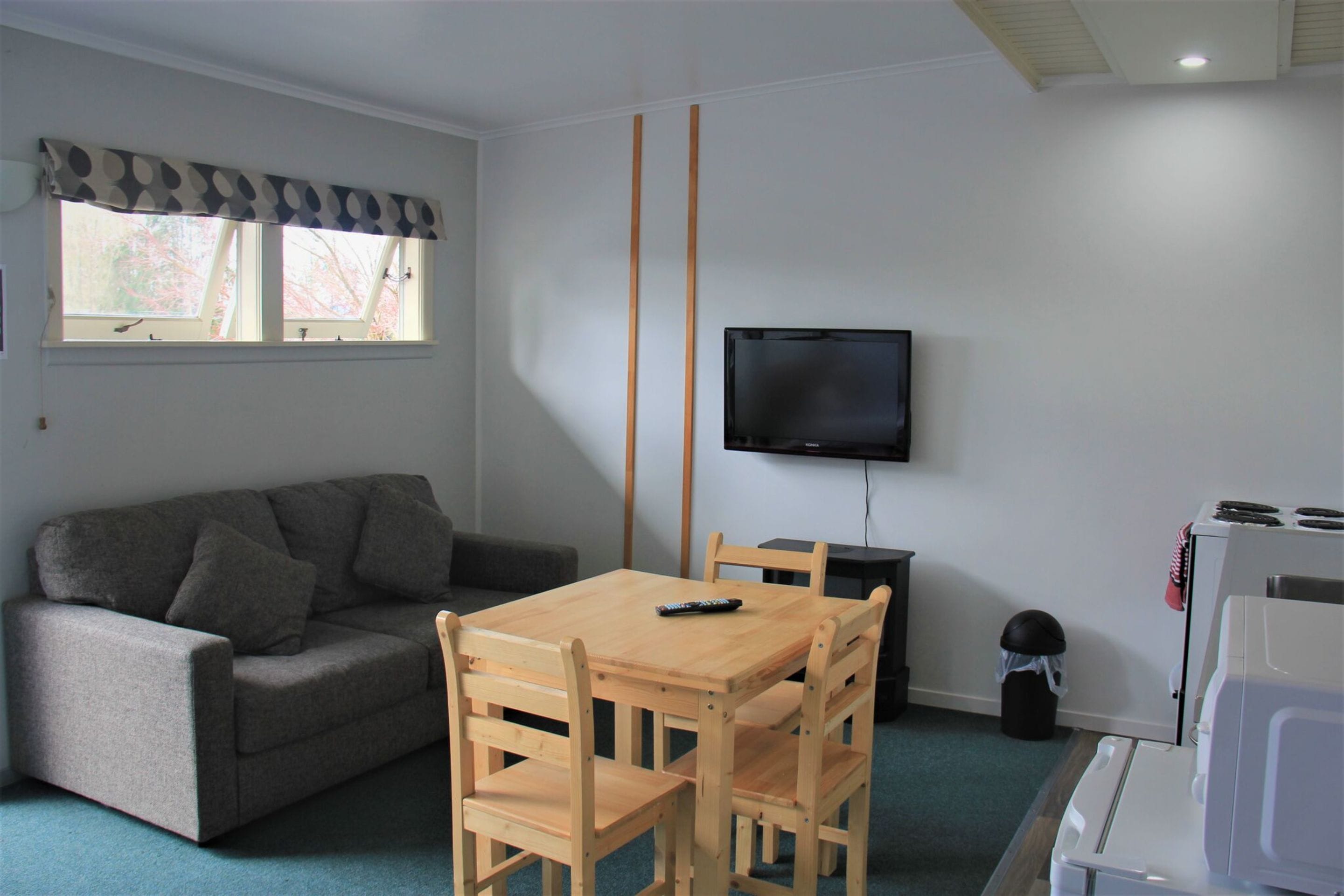 Un espace de vie compact comprenant un canapé, une table à manger en bois, des chaises, un téléviseur fixé au mur et un petit coin cuisine.