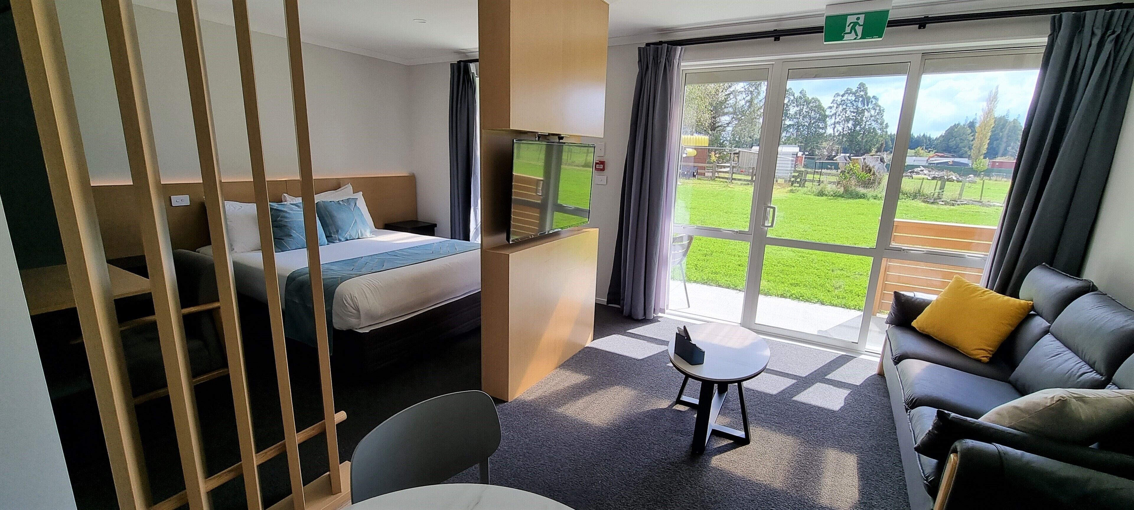 Ein Hotelzimmer mit einem Bett, einem Sofa, einem kleinen Tisch, einem Fernseher und einem großen Fenster mit Blick auf eine Grünfläche.