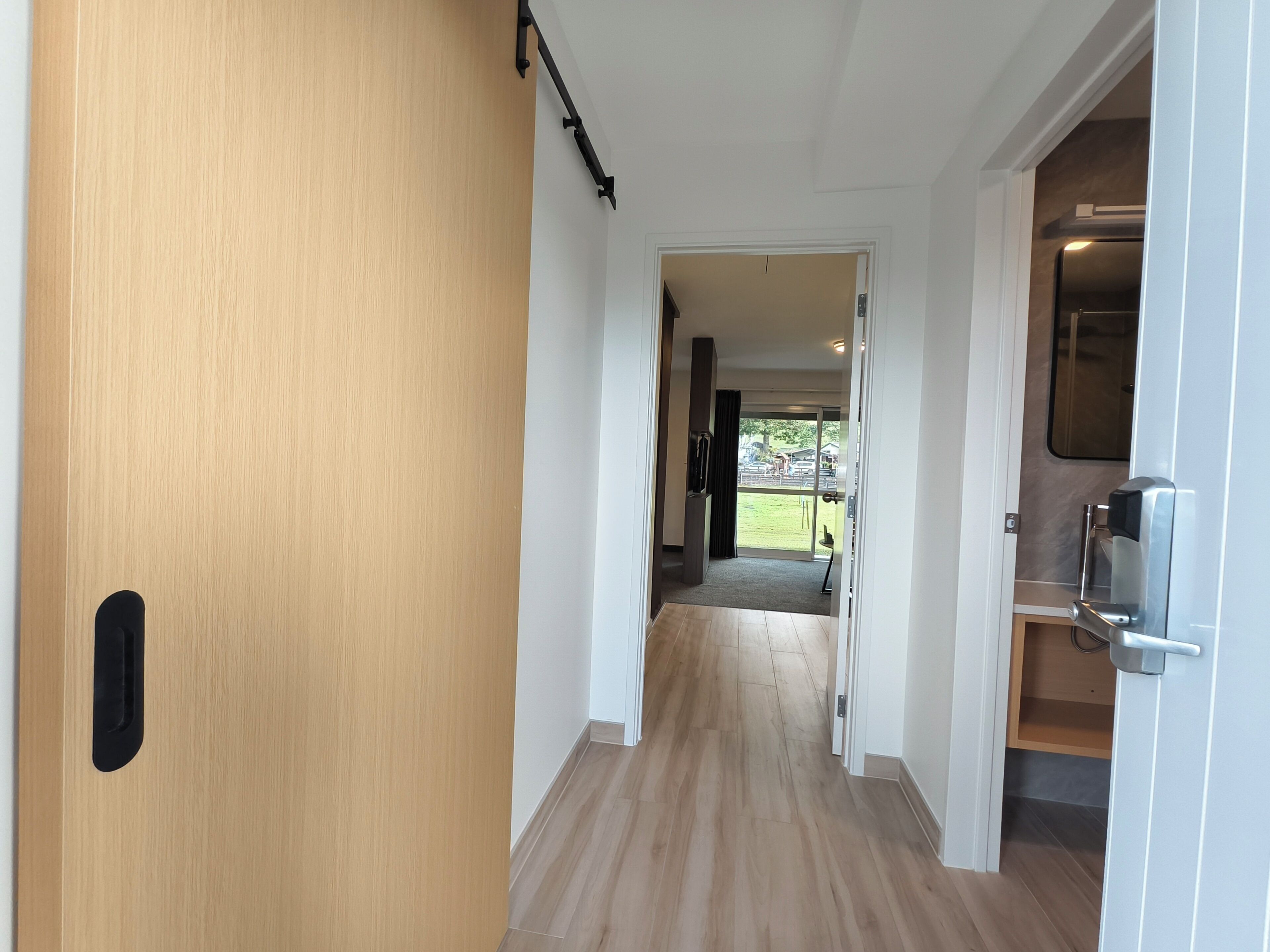 A hallway with a wooden door on the left, a white wall, and a door leading to a balcony with a view of the outside.
