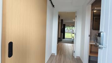 A hallway with a wooden door on the left, a white wall, and a door leading to a balcony with a view of the outside.