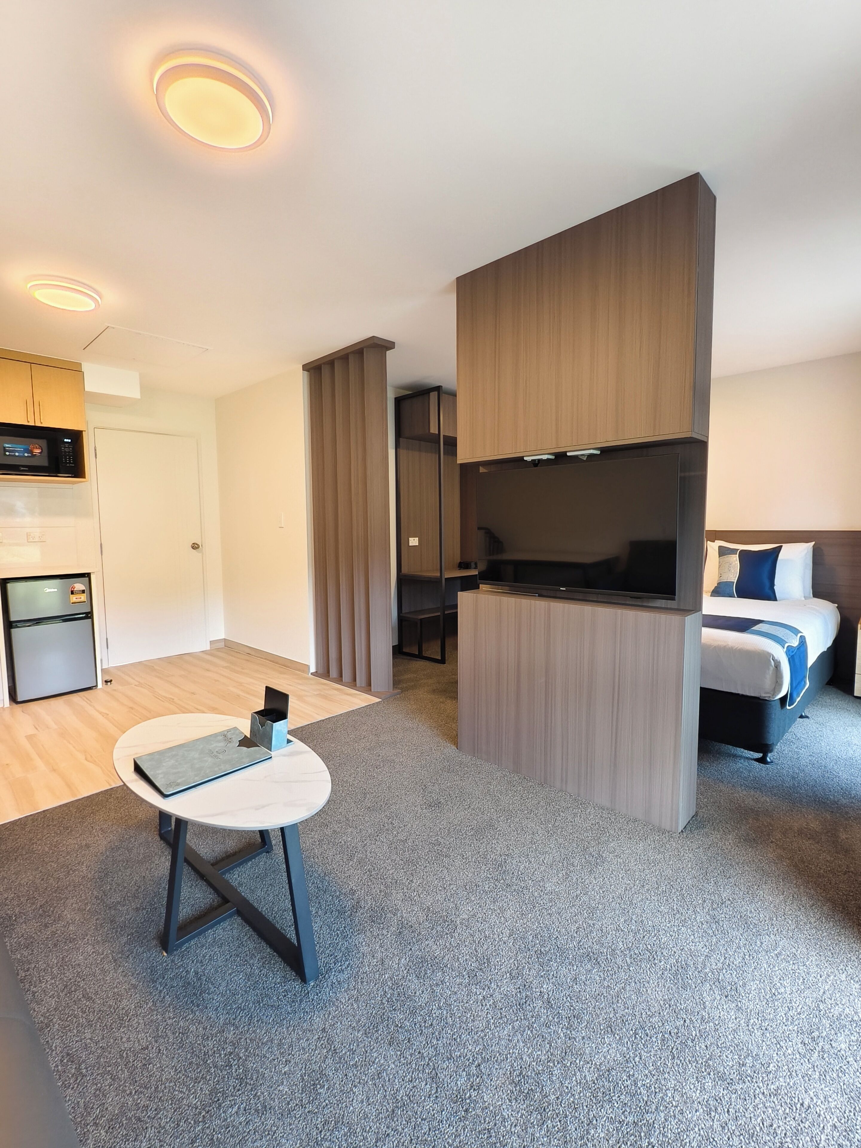 A modern hotel room with a kitchenette, a bed with blue bedding, a round table, and a grey carpet.