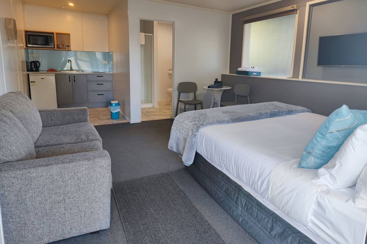 A compact hotel room with a bed, two armchairs, a small kitchenette, and a bathroom.