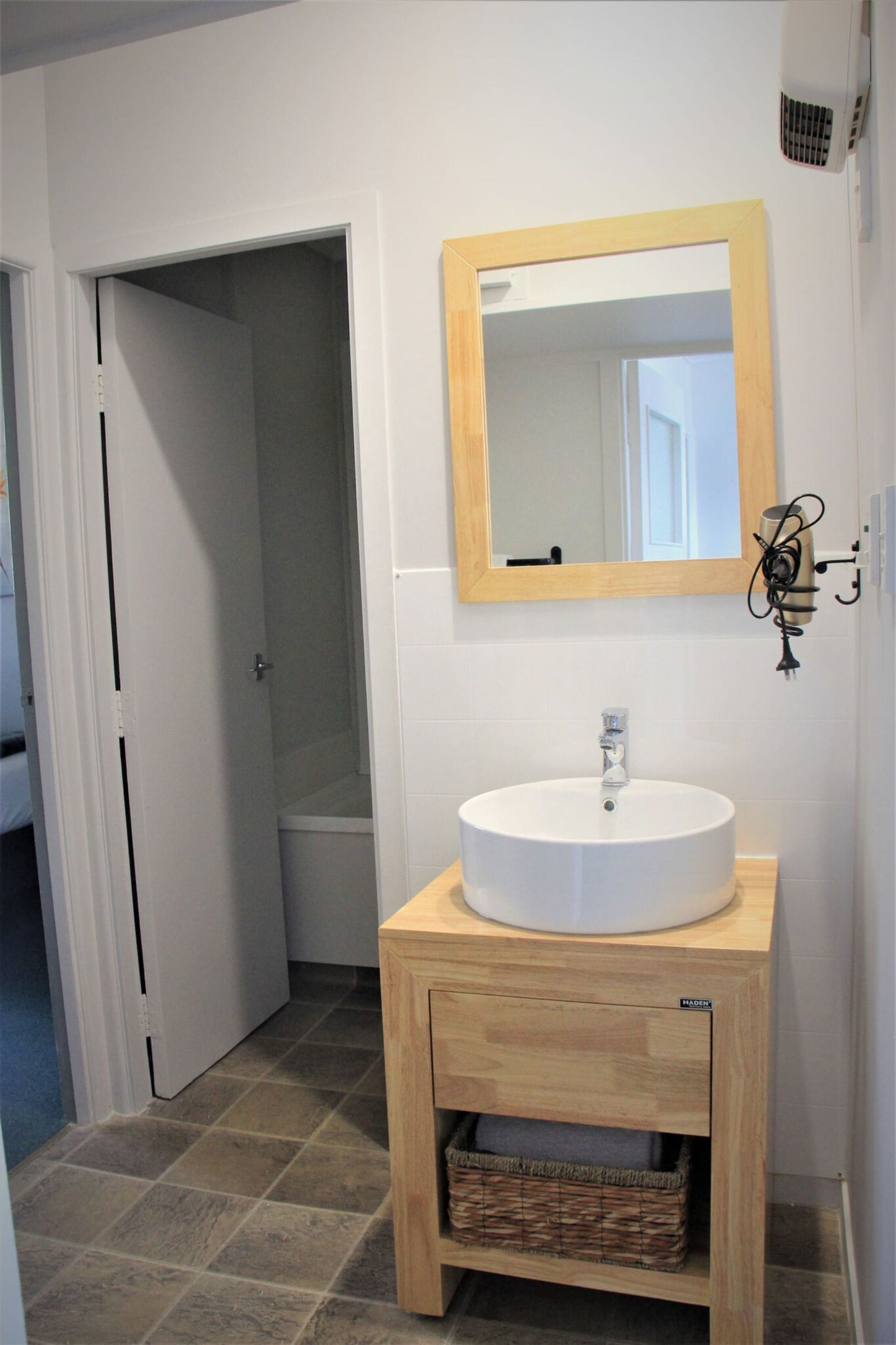 Une salle de bain avec un lavabo blanc, un meuble-lavabo en bois, un miroir et une porte donnant sur une autre pièce.