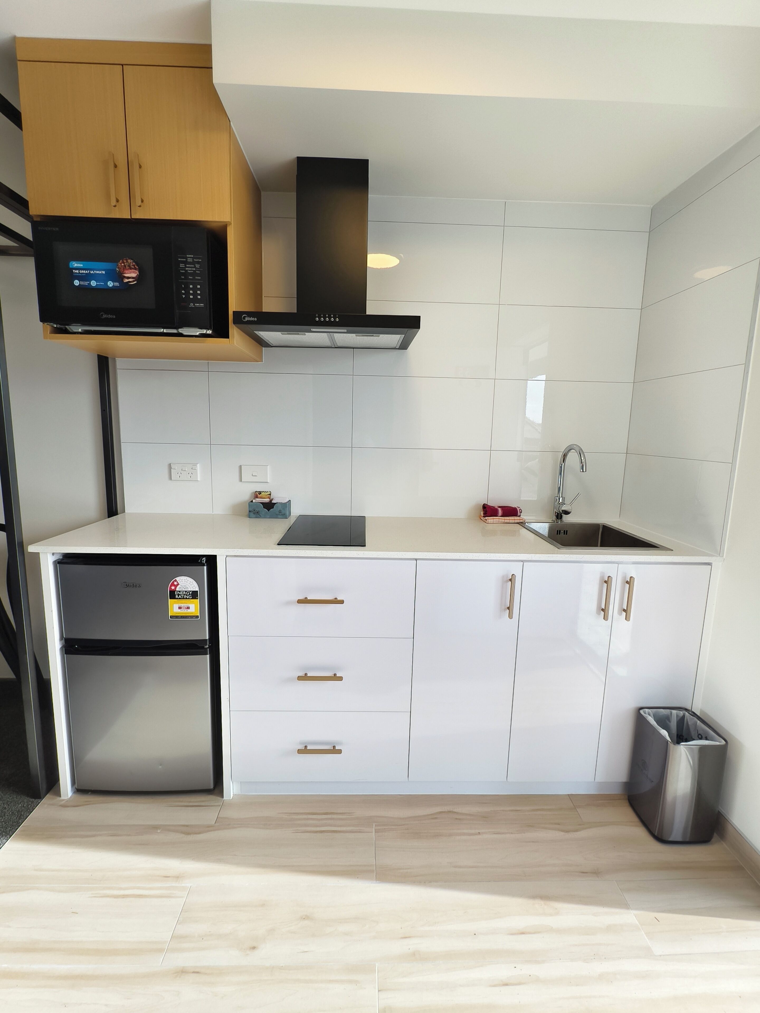 Una cocina moderna con armarios blancos, microondas integrado, pileta de acero inoxidable y refrigerador compacto.