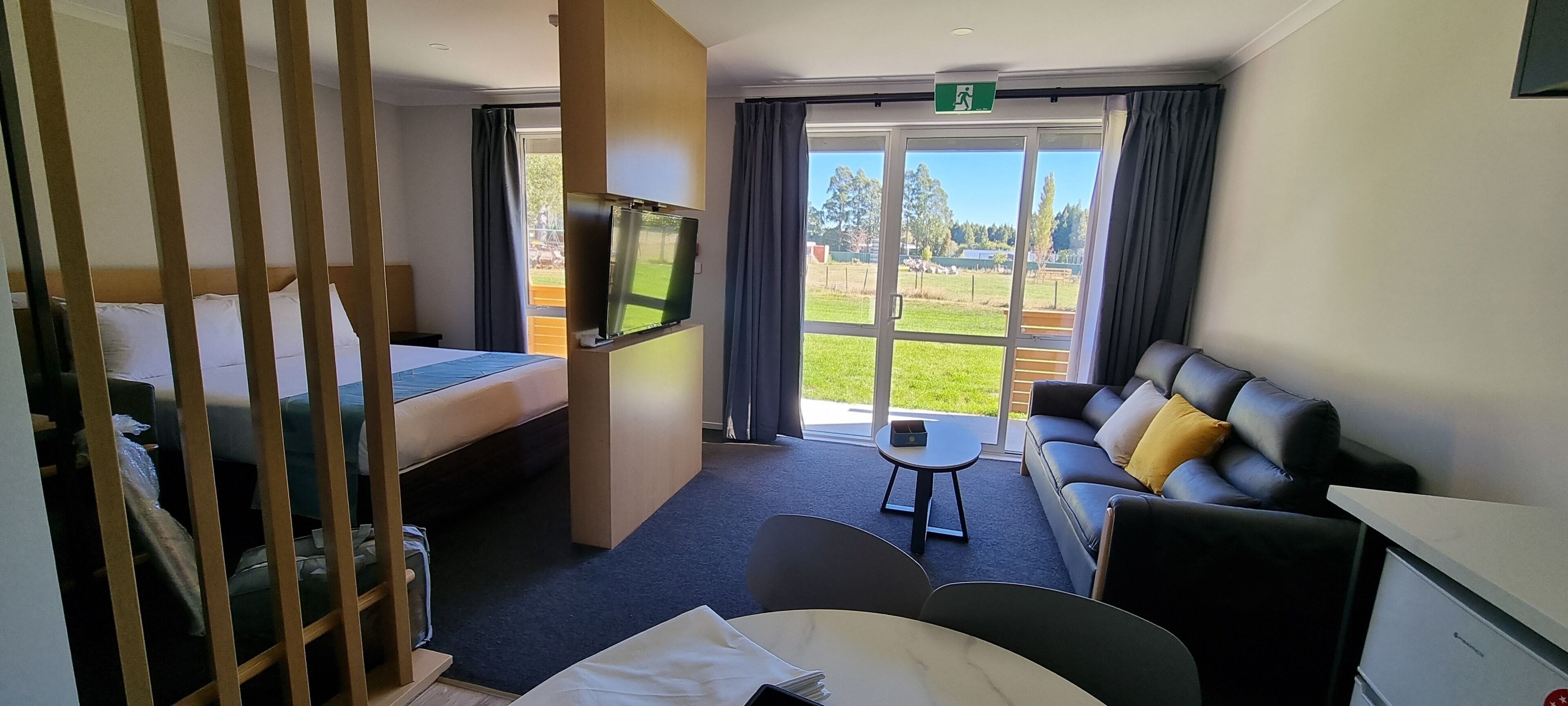 A hotel room with a bed, a sofa, a TV, a small table, and a view of a green landscape through large windows.