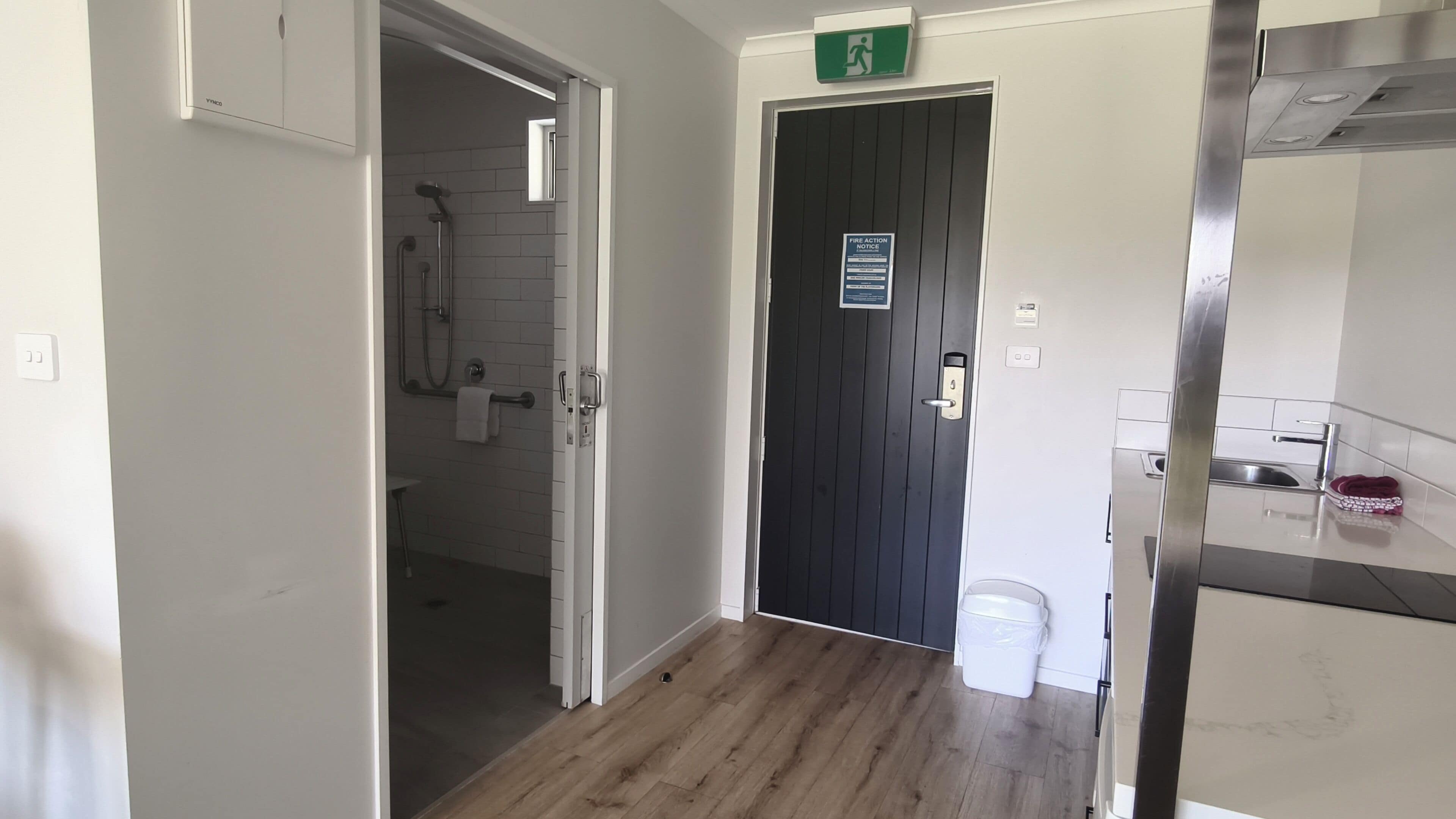A hallway leading to a bathroom with a shower, a door with a fire action notice, and a kitchen area with a sink and a trash bin.