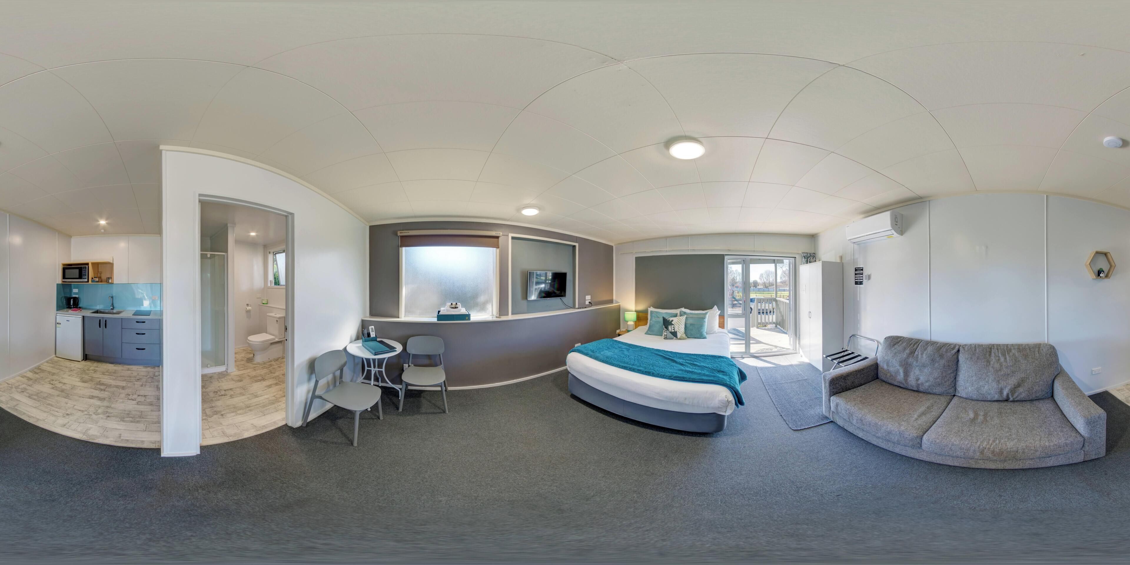 A modern hotel room with a curved bed, a sofa, a desk with a chair, a small table, and a bathroom with a shower and toilet.