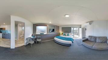A modern hotel room with a curved bed, a sofa, a desk with a chair, a small table, and a bathroom with a shower and toilet.