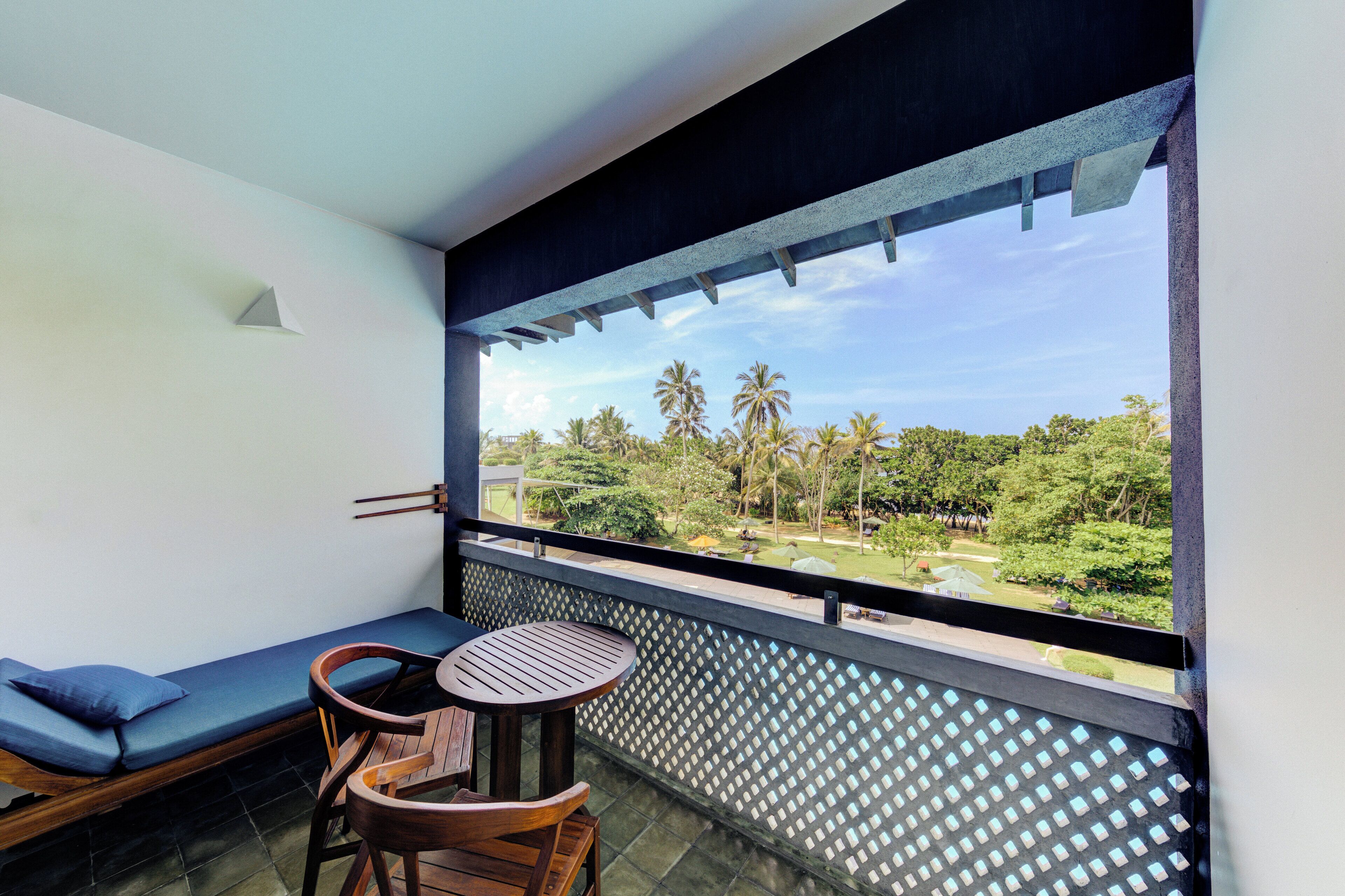 A balcony with a view of a garden, a seating area with a table and chairs, and a small window with a triangular design.