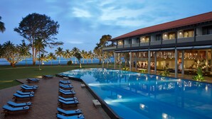 A pool with lounge chairs, a building with a red roof, and palm trees. - Cinnamon Bey Beruwala (Beruwala)
